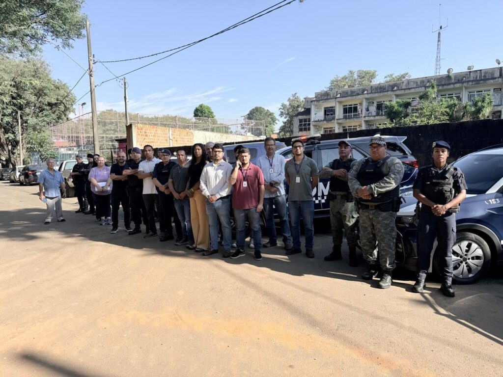 A imagem mostra um grupo de cerca de 20 pessoas alinhadas ao ar livre. Entre elas, há homens e mulheres, alguns vestindo uniformes que parecem ser de forças de segurança ou policiais, com coletes táticos, calças camufladas e camisas escuras. Outros estão com roupas civis, como camisas casuais e calças. Eles estão em um terreno de terra ou cascalho, e atrás deles há vários veículos escuros, possivelmente carros policiais, com algumas marcações visíveis, como o número "153" e a letra "N". Ao fundo, é possível ver edifícios com janelas e uma cerca, sugerindo que o local pode ser uma instituição ou uma instalação de segurança, talvez uma delegacia ou prisão. O céu está claro e azul, indicando que a foto foi tirada durante o dia. A maioria das pessoas olha para a câmera ou ligeiramente para o lado, com expressões neutras ou sérias. A foto é capturada na altura dos joelhos para cima, com foco no grupo e nos veículos atrás, transmitindo a ideia de uma reunião formal, operação conjunta ou uma foto de equipe.