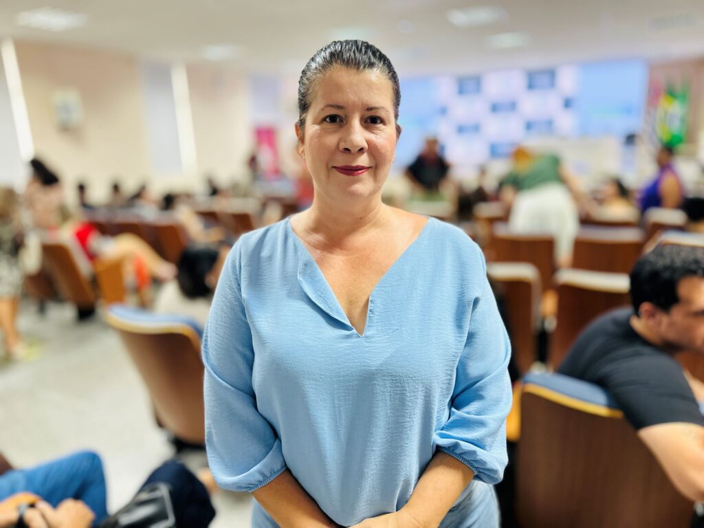 A imagem mostra uma mulher de meia-idade, com cabelos escuros presos em um coque, pele clara e uma expressão suave e amigável. Ela está vestindo uma blusa azul clara de manga três quartos com decote em V. A mulher está de pé, com as mãos entrelaçadas à frente do corpo, olhando diretamente para a câmera, transmitindo uma sensação de confiança e serenidade. O fundo é desfocado, revelando um auditório cheio de pessoas sentadas em cadeiras marrons e azuis, sugerindo que a mulher pode estar em um evento, palestra ou conferência. A iluminação é clara e uniforme, destacando bem a figura da mulher no centro da imagem. O ambiente transmite uma atmosfera profissional e acolhedora.