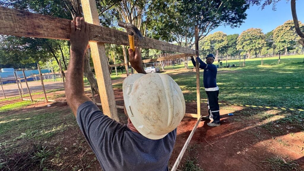 A imagem mostra dois homens trabalhando na construção de uma estrutura de madeira ao ar livre. Um dos homens, usando capacete branco, está segurando uma peça vertical de madeira no lugar, enquanto o outro, com capacete azul escuro e roupas de trabalho, está martelando um prego em uma viga horizontal de madeira. O ambiente ao redor é um campo gramado com algumas árvores ao fundo e uma cerca azul, possivelmente de uma quadra esportiva, ao longe. O chão é de terra com algumas áreas de grama. A luz do sol é forte, indicando que é durante o dia, provavelmente no fim da tarde, criando sombras e destacando as texturas rústicas da madeira e do solo. A cena transmite uma sensação de trabalho manual e construção em andamento, possivelmente em um parque ou área de lazer.