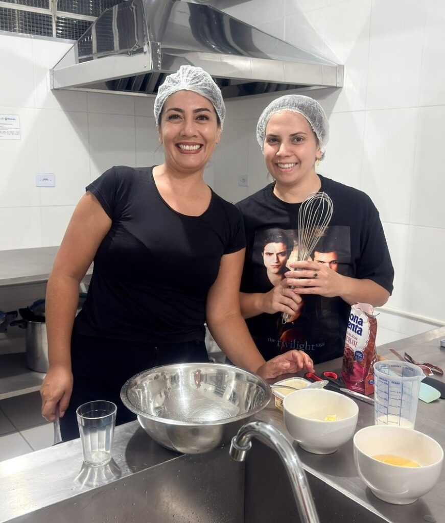 A imagem mostra duas mulheres em uma cozinha, aparentemente preparando uma receita de bolo ou sobremesa. Ambas usam toucas para cabelo, provavelmente por higiene alimentar, e estão sorrindo para a câmera, transmitindo uma sensação de alegria e colaboração. À esquerda, a mulher veste uma camiseta preta e calça escura, enquanto a mulher à direita usa uma camiseta preta com uma estampa gráfica e segura um batedor manual (fouet). Na bancada à frente delas, há vários utensílios e ingredientes dispostos: uma tigela grande de metal, algumas tigelas menores contendo manteiga, ovos e outros ingredientes, um copo com líquido e uma embalagem de mix para chocolate da marca "FONTANA". Também é possível ver uma pia de aço inoxidável com uma torneira, e ao fundo, uma coifa de inox sobre o fogão, além de paredes revestidas com azulejos brancos. A iluminação é clara e uniforme, destacando os detalhes dos ingredientes e utensílios, e o ambiente transmite uma atmosfera organizada e acolhedora, típica de uma cozinha profissional ou de uma aula de culinária.