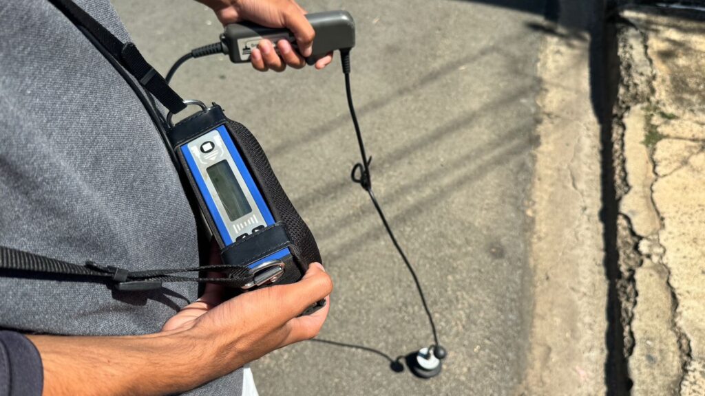 A imagem mostra uma pessoa segurando um dispositivo eletrônico portátil, que parece ser um instrumento de medição ou um equipamento técnico. O dispositivo é azul e preto, possui uma tela digital e alguns botões. A pessoa segura também um componente cinza conectado ao dispositivo principal por um cabo. Na outra extremidade do cabo, há um sensor pequeno e redondo com duas protuberâncias esféricas. A pessoa está usando uma alça preta para segurar o equipamento, que passa sobre o ombro. O ambiente parece ser externo, possivelmente uma calçada ou rua, com o chão de concreto e uma guia visível à direita. A iluminação indica que é durante o dia, com sombras projetadas no chão. A foto está focada no equipamento e nas mãos da pessoa, mostrando detalhes claros dos dispositivos e do entorno imediato.