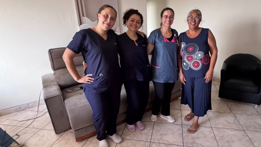 A imagem mostra quatro mulheres posando juntas em um ambiente interno, possivelmente uma sala de estar ou uma área comum. Três delas estão vestindo uniformes médicos azuis, sugerindo que são profissionais de saúde. A mulher mais à direita é mais velha e veste uma blusa azul-marinho sem mangas com um padrão circular em tons de rosa e branco, além de calças azuis. Todas estão sorrindo, transmitindo uma sensação de amizade e companheirismo. Ao fundo, há um sofá cinza reclinável e outra poltrona preta, com piso de cerâmica clara. A iluminação é boa e natural, indicando que a foto foi tirada durante o dia.