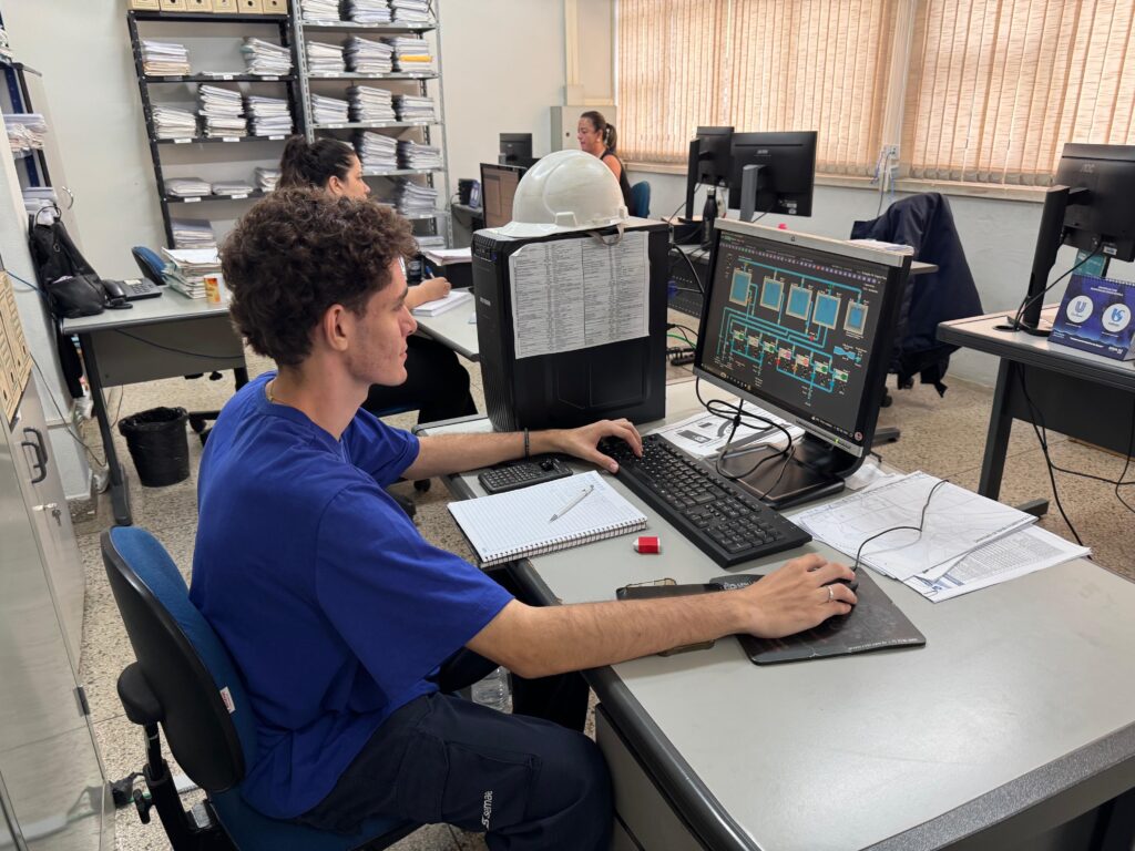A imagem mostra um ambiente de escritório ou sala de controle, onde um jovem está sentado em frente a um computador. Ele usa uma camiseta azul com o logo "Semae" na calça e está focado na tela do computador, que exibe um diagrama técnico, provavelmente de um sistema de tratamento de água, com tanques e tubulações representados em azul. Sobre o gabinete do computador há um capacete branco, sugerindo um ambiente industrial ou técnico. Na mesa, há um caderno aberto com uma caneta e uma calculadora, indicando que ele pode estar realizando alguma anotação ou cálculo relacionado ao trabalho. Ao fundo, é possível ver outras duas pessoas trabalhando e estantes metálicas cheias de pastas e documentos. O ambiente é bem iluminado, com janelas grandes e persianas verticais que deixam entrar a luz do dia. A imagem transmite um ambiente profissional e técnico, possivelmente ligado à engenharia ou ao controle de processos industriais, especialmente na área de tratamento de água, conforme indicam os termos visíveis na tela do computador.