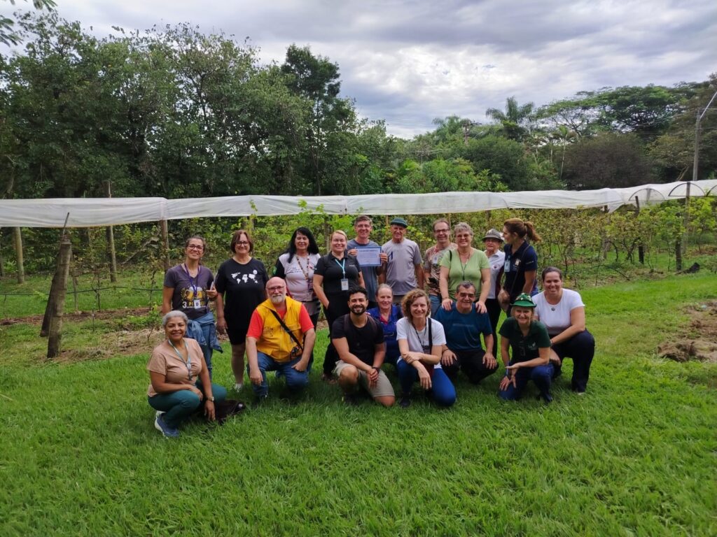 A imagem mostra um grupo de cerca de 17 pessoas posando para uma foto em um ambiente agrícola ao ar livre, provavelmente uma vinha. O grupo está organizado em duas fileiras: algumas pessoas agachadas ou ajoelhadas na frente e outras em pé atrás. Todos estão vestidos de forma casual, com roupas adequadas para atividades ao ar livre, como camisetas, calças jeans e chapéus. Um homem no centro da fileira de trás segura um certificado azul e branco, sugerindo que o grupo pode estar comemorando a conclusão de um curso, treinamento ou algum tipo de reconhecimento. Ao fundo, é possível ver fileiras de videiras cobertas por uma proteção branca, possivelmente uma rede ou plástico para proteger as plantas. Mais atrás, há uma área com muitas árvores verdes e o céu está nublado, com nuvens acinzentadas que criam uma iluminação suave e difusa na cena. O chão em frente ao grupo é uma grama verde bem cuidada. A atmosfera geral da foto é alegre e de confraternização, em meio a um cenário natural e agrícola.