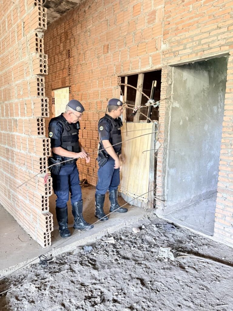 A imagem mostra dois homens vestidos com uniformes que lembram os de policiais ou agentes de segurança, dentro de um prédio em construção ou abandonado. As paredes são de tijolos aparentes e o chão está sujo, com detritos e poeira. Um dos homens está olhando para baixo, enquanto o outro olha para uma abertura que parece ser uma porta ou um corredor. Ambos usam botas pretas e bonés, e seus uniformes são azuis escuros com distintivos. A iluminação é natural, mas o ambiente está um pouco escuro, sugerindo que a foto foi tirada durante o dia, porém em um local sem muita luz direta. O cenário transmite uma sensação de seriedade, possivelmente uma inspeção ou investigação em andamento.