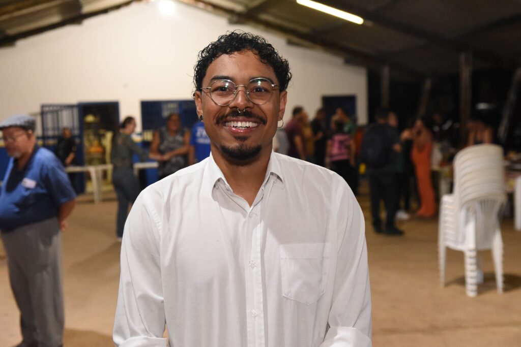 A imagem mostra um jovem com cabelo encaracolado, óculos e um piercing no septo do nariz. Ele está sorrindo amplamente e olhando diretamente para a câmera. Ele veste uma camisa branca com gola. O fundo está desfocado, mas é possível perceber que se trata de um ambiente interno, possivelmente uma reunião ou evento social, com outras pessoas e mesas ao fundo, além de algumas cadeiras brancas empilhadas. A iluminação é artificial, vinda de luzes no teto, e destaca o rosto do jovem, transmitindo uma sensação de simpatia e acessibilidade.