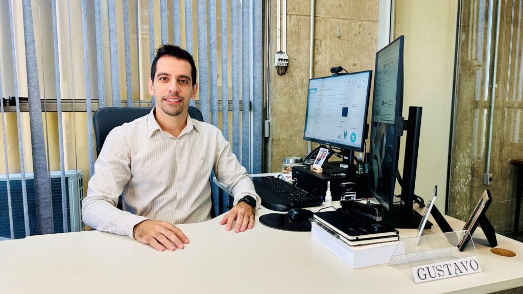 A imagem mostra um homem sentado em uma mesa de escritório branca em formato de L. Ele aparenta estar na faixa dos 30 anos, tem cabelo curto e escuro, além de barba por fazer. Está vestindo uma camisa clara com listras verticais e usa um relógio preto no pulso esquerdo. O homem está olhando diretamente para a câmera com um leve sorriso. Na mesa, há dois monitores de computador: um na horizontal exibindo gráficos e dados, e outro na vertical mostrando uma lista ou feed. Também estão presentes um teclado preto, um mouse com mousepad, um grampeador e alguns cadernos empilhados sobre papéis brancos. Um pequeno porta-nome com o nome "GUSTAVO" está visível na mesa. Ao fundo, há uma janela grande com persianas verticais azuis, que permitem a entrada de luz natural. A parede ao lado é de concreto aparente, com canaletas elétricas expostas e uma pequena caixa elétrica branca. A iluminação é clara e uniforme, dando um ambiente profissional e organizado ao escritório.