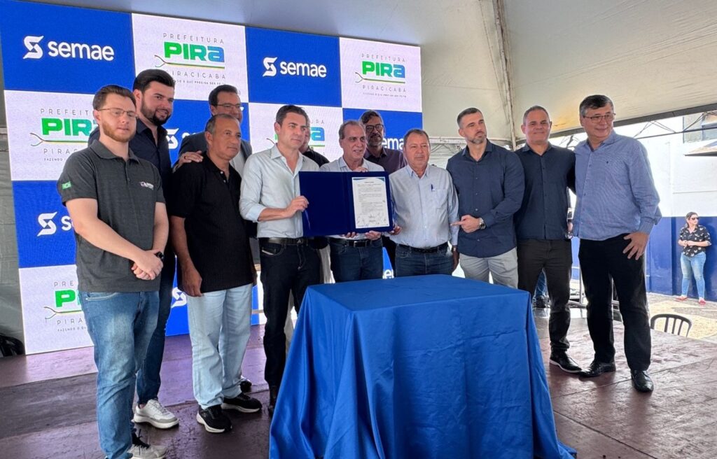 A imagem mostra um grupo de dez homens alinhados atrás de uma mesa coberta por uma toalha azul, em um ambiente externo, possivelmente durante um evento oficial ou cerimônia. Eles estão vestidos de forma casual social, com camisas, polos e calças jeans. No centro do grupo, um homem segura um documento em uma pasta azul, sugerindo a assinatura ou apresentação de um acordo ou certificado. Ao fundo, há uma grande faixa com padrão quadriculado azul e branco, contendo os logotipos e nomes "PREFEITURA", "PIRA", "PIRACICABA" e "SEMAE", indicando que o evento está relacionado à Prefeitura de Piracicaba e ao Serviço Municipal de Água e Esgoto (SEMAE). A iluminação é natural, típica de um evento ao ar livre durante o dia. No canto direito da imagem, é possível ver uma mulher parcialmente, que parece estar observando a cerimônia. A composição transmite um momento formal e comemorativo, com os participantes aparentando estar satisfeitos ou orgulhosos.
