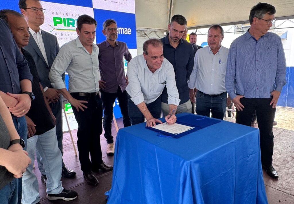 A imagem mostra uma cerimônia oficial de assinatura de um documento. No centro, um homem está inclinado sobre uma mesa azul, assinando um papel. Ele veste uma camisa azul clara. Ao seu redor, há outros cinco homens, todos observando atentamente o ato da assinatura. Eles estão vestidos de forma formal, com camisas sociais, algumas de manga longa e outras de manga curta. A mesa está coberta por um tecido azul vibrante, e sobre ela há pastas e documentos. Ao fundo, há um banner com as palavras "PREFEITURA PIRACICABA" e o slogan "FAZENDO O QUE É PRECISO SER FEITO", indicando que o evento está relacionado a uma ação oficial da prefeitura da cidade de Piracicaba. O ambiente parece ser ao ar livre ou em um local muito bem iluminado, com piso de pedras ou concreto. A expressão dos homens é séria e concentrada, evidenciando a importância do momento.