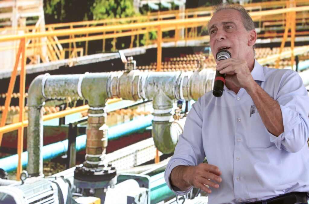 A imagem mostra um homem de meia-idade falando ao microfone. Ele está vestido com uma camisa azul clara de manga comprida, com as mangas dobradas até o cotovelo, e parece estar em meio a uma apresentação ou discurso. O homem segura o microfone com a mão direita, enquanto a mão esquerda está levantada, fazendo um gesto, possivelmente para enfatizar algum ponto. Ele tem cabelo curto e está olhando ligeiramente para cima e para a esquerda, com a boca aberta, indicando que está falando. Ao fundo, há uma imagem desfocada e colorida que parece representar uma estrutura industrial, com tubulações e elementos metálicos em tons de amarelo, verde e azul. A iluminação destaca o rosto do homem, criando um contraste com o fundo mais escuro e desfocado. A composição sugere que o foco principal é o homem e seu discurso, enquanto o fundo fornece um contexto visual relacionado ao tema da apresentação, possivelmente ligado a indústria ou infraestrutura.