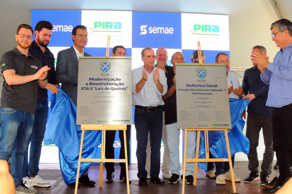 A imagem mostra uma cerimônia oficial de inauguração ou entrega de obras públicas na cidade de Piracicaba, Brasil. No centro da imagem, há duas placas comemorativas grandes, montadas em cavaletes de madeira, que estão sendo descerradas por um grupo de homens. As placas contêm textos que se referem a projetos de modernização e reforma relacionados ao sistema de água e esgoto, especificamente à Estação de Tratamento de Água (ETA) II "Luiz de Queiroz" e à Estação Elevatória da Captação do Rio Piracicaba. Ao redor das placas, há cerca de dez homens, a maioria vestindo roupas formais ou sociais, como camisas, calças e alguns com ternos. Eles parecem ser autoridades municipais e representantes do Serviço Municipal de Água e Esgoto (SEMAE), já que os nomes e cargos aparecem nas placas e nos banners ao fundo. Alguns estão aplaudindo, outros seguram as cortinas azuis usadas para cobrir as placas antes da revelação. No fundo, banners azuis e brancos exibem os logos da Prefeitura de Piracicaba e do Semae, reforçando o caráter oficial do evento. A iluminação é clara e destaca bem os participantes e as placas, evidenciando a importância da ocasião. A cerimônia parece celebrar a entrega ou início de melhorias em infraestrutura hídrica da cidade, com menção a autoridades como o prefeito Helio Donizete Zanatta e o vice-prefeito Sergio José Dias Pacheco Junior. A data indicada nas placas é abril de 2026, sugerindo que o evento é recente ou futuro próximo. Em resumo, a foto registra um momento solene e simbólico de avanço em serviços públicos essenciais para a população de Piracicaba, com a presença de autoridades locais e representantes do setor de saneamento.