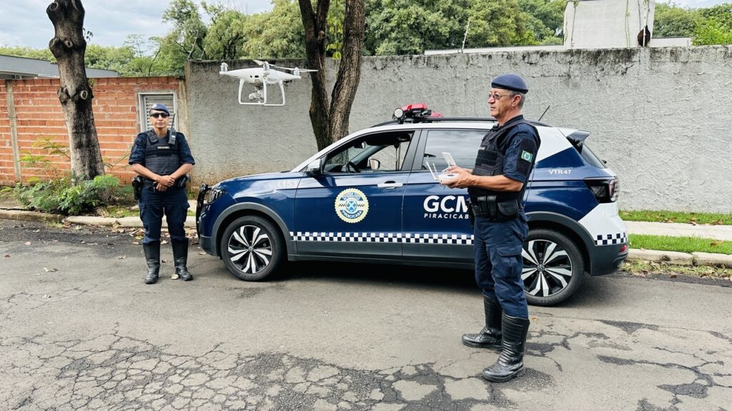 A imagem mostra dois agentes da Guarda Civil Municipal (GCM) de Piracicaba, Brasil, em uma ação com um drone. Eles estão posicionados ao lado de uma viatura da GCM, que é um veículo SUV de cor escura com os símbolos e inscrições da corporação, incluindo "GCM PIRACICABA" e a placa "VTR47". Um dos agentes está operando o controle do drone, que está voando logo à frente deles, enquanto o outro está em posição de atenção, observando o drone. Ambos vestem uniformes azul escuro com coletes táticos, boinas e botas pretas. O cenário é ao ar livre, com uma parede de tijolos ao fundo à esquerda, uma parede de concreto à direita e algumas árvores ao fundo, sugerindo uma área urbana ou suburbana. A iluminação é natural, indicando que a foto foi tirada durante o dia. A cena transmite uma ação de fiscalização ou monitoramento pela GCM utilizando tecnologia de drones para reforçar a segurança pública.