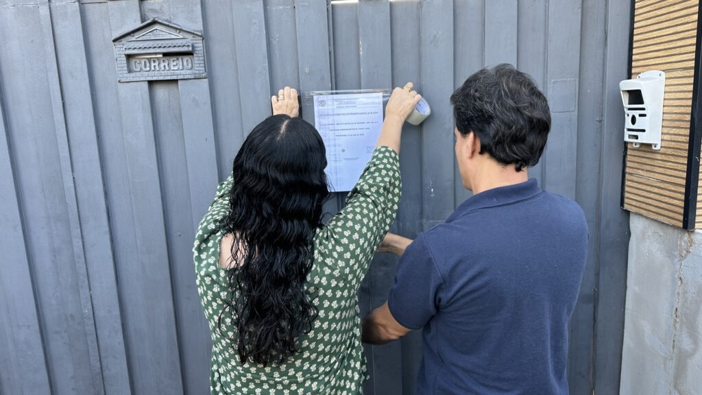 A imagem mostra uma mulher de cabelos pretos longos e blusa verde com detalhes branco, cola um aviso em porta de ferro de estabelecimento, observada por um homem de camiseta azul escura, que ajuda a segurar o folha.