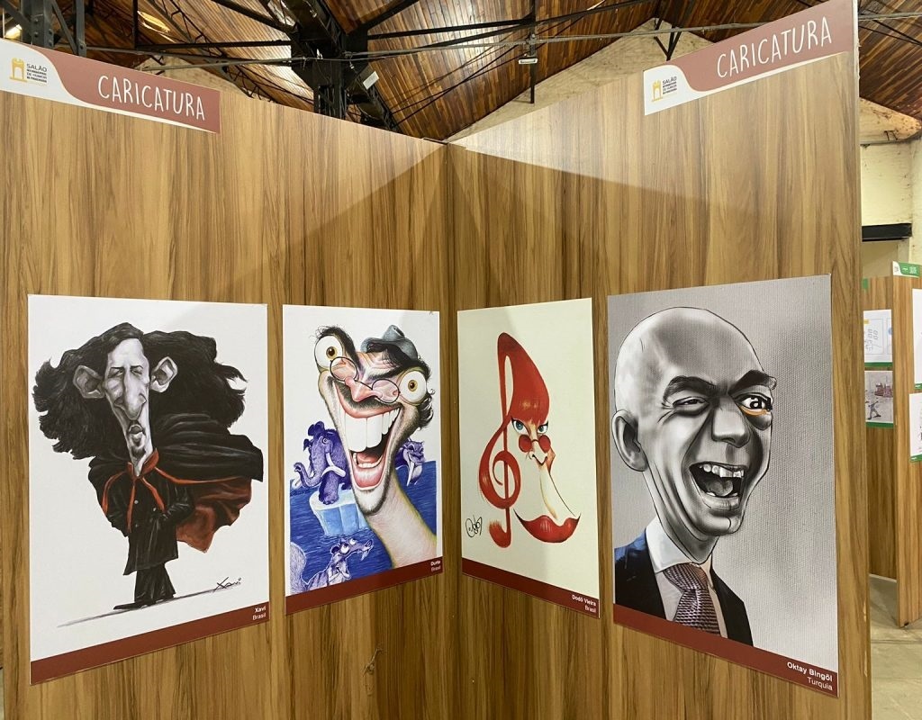 A imagem mostra um canto de uma exposição de caricaturas em um ambiente interno com paredes revestidas em madeira clara e teto com vigas metálicas escuras e ripas de madeira. Na parede da esquerda, há duas caricaturas coloridas: a primeira retrata um homem com cabelos longos e escuros, vestindo uma capa, assinada por "Xavi Brasil"; a segunda mostra um homem de olhos arregalados e sorriso largo, com mamutes ao fundo, assinada por "Duarte Brasil". Na parede da direita, também há duas caricaturas: uma com um desenho criativo onde uma clave de sol vermelha forma o perfil de um rosto com óculos e lábios vermelhos, assinada por "Dodô Vieira Brasil"; e outra em preto e branco de um homem careca rindo, usando terno e gravata, assinada por "Oktay Bingöl Turquia". Acima das caricaturas, placas marrons com letras brancas indicam o tema "CARICATURA" e o nome do evento "Salão Internacional de Humor de Piracicaba". A iluminação é quente e destaca as obras, criando um ambiente acolhedor para apreciar as caricaturas.
