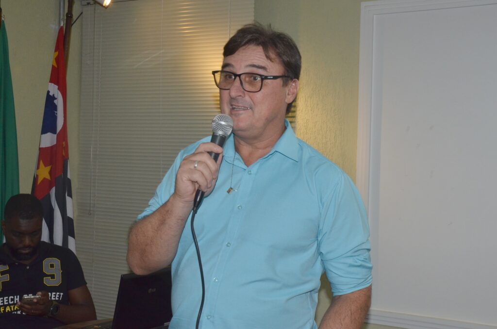 A imagem mostra um homem de meia-idade falando em um microfone, aparentemente durante uma apresentação ou reunião. Ele usa óculos, uma camisa azul clara e um colar com um pingente quadrado. O homem está com uma expressão concentrada, olhando para frente enquanto segura o microfone com a mão direita. À esquerda da imagem, há outro homem sentado, que parece estar olhando para o celular ou para algo em suas mãos. Ele veste uma camisa preta com detalhes em amarelo e branco e está próximo a um laptop. Ao fundo, há uma parede com textura e uma janela com persianas fechadas. Também é possível ver parcialmente uma bandeira com cores vermelho, branco, azul, preto e amarelo, que está posicionada atrás dos homens. A iluminação é quente e cria sombras no ambiente, sugerindo que o local pode ser uma sala de conferências ou reunião.