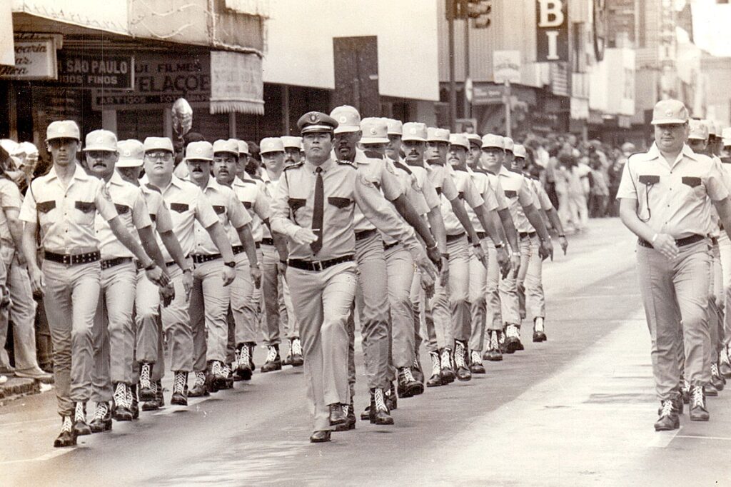 A imagem mostra um grupo de homens marchando em formação, provavelmente em um desfile ou cerimônia oficial. Eles vestem uniformes compostos por camisas claras de manga curta, gravatas escuras, calças escuras e botas pretas com cadarços brancos. Todos usam chapéus e cintos escuros, e alguns parecem usar relógios. A marcha é sincronizada, com expressões sérias e focadas. O cenário é uma rua urbana, com edifícios comerciais ao fundo, onde se podem ver algumas placas com inscrições em português, como "SÃO PAULO", "FILMES" e "ARTIGOS PARA", sugerindo que a cena ocorre em uma cidade brasileira, possivelmente São Paulo. A fotografia é em preto e branco, com boa iluminação natural, e o foco está nos homens em primeiro plano, enquanto o fundo está desfocado. A imagem transmite uma sensação de disciplina, ordem e formalidade.