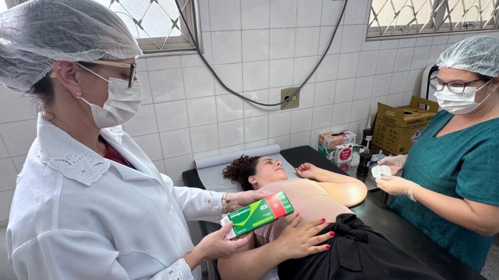 A imagem mostra um ambiente clínico onde três pessoas estão presentes. À esquerda, um profissional de saúde usando jaleco branco, touca, óculos e máscara segura uma caixa de medicamento verde e vermelha, na qual se lê "etonogestrel" e "136". No centro, uma paciente está deitada em uma maca, vestindo uma blusa clara e calça preta com um laço na cintura, olhando para cima. À direita, outro profissional de saúde, também com touca e máscara, está segurando um curativo branco e luvas, aparentemente preparando-se para realizar um procedimento. O ambiente é iluminado artificialmente, com paredes revestidas de azulejos claros e uma janela com grades em forma de losango. Ao fundo, há caixas e frascos de materiais médicos. A cena transmite uma situação de atendimento médico, possivelmente relacionada à administração do medicamento mencionado.