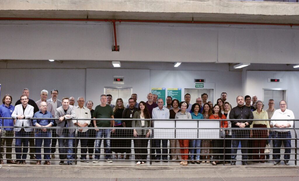 A imagem mostra um grande grupo de pessoas reunidas para uma foto em um ambiente interno, possivelmente um prédio comercial ou corporativo. O grupo está posicionado atrás de uma grade ou corrimão metálico, em um espaço que parece ser um mezanino ou uma área elevada. As pessoas estão alinhadas em uma única fila, olhando para a câmera, com expressões neutras ou de leve sorriso. O local tem um estilo arquitetônico moderno, com paredes de concreto aparente e um teto com iluminação embutida. Ao fundo, é possível ver portas de elevador e algumas placas indicativas de andares e setores, como "11º andar", "4º andar" e "1º andar". Também há placas que indicam setores da empresa, como "Comercial", "Financiamento", "Serviços Financeiros", "Serviços Administrativos", "Ações de Marketing" e "Rede de Vendas". Uma placa de segurança orienta, em caso de acidente, avisar o seguro. A iluminação é artificial, clara e uniforme, destacando o grupo e o ambiente ao redor. A composição da imagem é formal, típica de uma foto institucional ou de equipe em um ambiente corporativo.