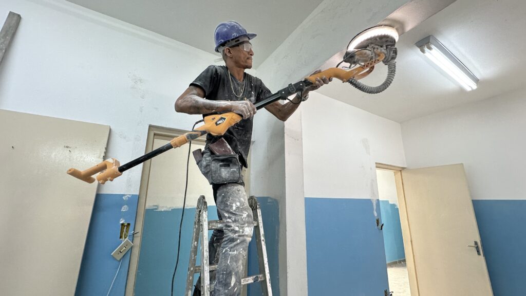 A imagem mostra um homem trabalhando na reforma de um ambiente, lustrando ou lixando o teto com uma lixa elétrica conectada a um cabo longo. Ele está em uma escada de alumínio e usa equipamentos de segurança, como capacete azul e óculos de proteção. Sua roupa está bastante suja de pó branco, provavelmente de gesso ou massa corrida. O ambiente tem as paredes pintadas na metade inferior de azul e a metade superior branca. A iluminação é forte, com luzes fluorescentes no teto e uma luz própria na lixa elétrica que ele está usando. O homem parece concentrado na tarefa, e ao fundo é possível ver uma porta aberta e um interruptor de luz pendurado na parede. A imagem transmite a ideia de um trabalho pesado e detalhado em reforma ou construção.