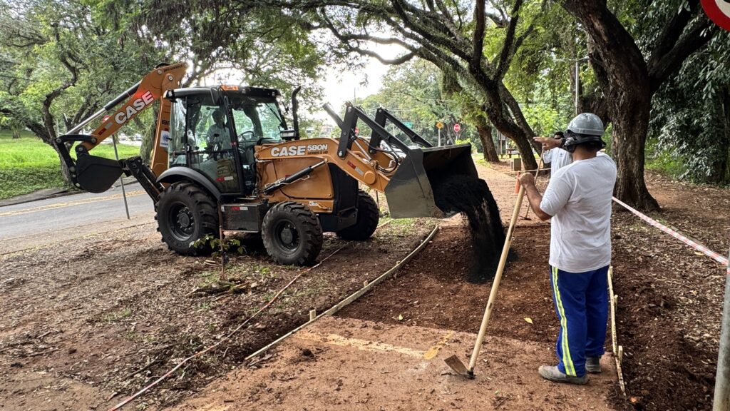 A imagem mostra uma cena de construção ao ar livre, provavelmente em um parque ou ao longo de uma estrada com muitas árvores. No centro da imagem, há uma retroescavadeira da marca CASE, modelo 580N Série 2 HD, de cor laranja vibrante, que está despejando terra do seu balde frontal. Ao lado da máquina, um trabalhador vestido com camiseta branca, calça azul com faixas amarelas e capacete branco está usando uma pá para ajudar no trabalho, possivelmente nivelando ou distribuindo a terra. O ambiente é cercado por árvores verdes e frondosas, com luz natural do dia filtrando pelas folhas, criando sombras e pontos de luz no chão. Há cones de sinalização laranja e fitas vermelhas e brancas delimitando a área de trabalho, indicando que é uma zona de construção ou manutenção. À esquerda, pode-se ver parte de uma estrada com faixas amarelas pintadas no asfalto. A cena transmite uma sensação de trabalho em andamento, com o maquinário pesado e o trabalhador colaborando na preparação do terreno, possivelmente para pavimentação ou jardinagem. A combinação da natureza ao redor com a atividade industrial cria um contraste interessante entre o ambiente natural e a intervenção humana.
