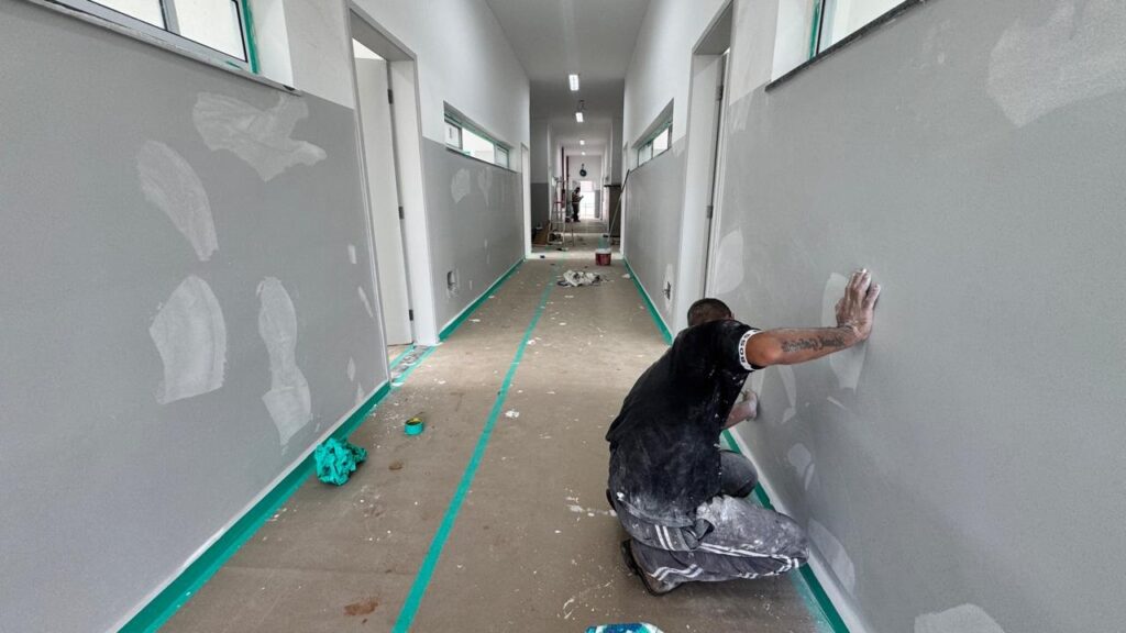 A imagem mostra um corredor longo e estreito em um ambiente interno que está em processo de construção ou reforma. Um homem, vestido com camiseta preta e calça de trabalho cinza, está agachado próximo à parede direita, com a mão direita encostada nela. A parede é pintada de cinza na parte inferior e branca na parte superior, apresentando várias manchas brancas de massa corrida ou gesso, indicando reparos. No braço direito do homem, é visível uma tatuagem, e na manga da camiseta está escrito "BOSS". O piso é de concreto, sujo de poeira, com duas linhas paralelas de fita adesiva verde fluorescente que percorrem todo o corredor, criando um efeito de profundidade na imagem. No fundo, é possível ver outras portas e janelas horizontais, além de outra pessoa ao longe. O ambiente está bem iluminado, provavelmente por luzes fluorescentes no teto. No chão, há alguns materiais e objetos de trabalho, como uma toalha verde e uma fita adesiva.