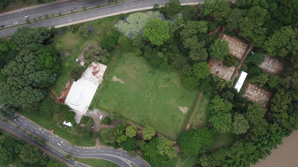 A imagem mostra uma vista aérea de um parque ou área verde com um campo esportivo retangular, provavelmente um campo de futebol, com gols visíveis nas extremidades. Ao lado do campo, há um grande edifício com uma estrutura de teto curva branca, que parece estar em construção ou reforma, com partes expostas e manchas avermelhadas. Ao lado do edifício e do campo, passa uma estrada com vários carros. Na parte esquerda da imagem, há um corpo d'água com folhas de lírio d'água. Do lado direito, uma área densa de árvores e vegetação cobre grande parte da cena, onde também se encontram algumas estruturas retangulares pavimentadas que parecem ser quadras antigas ou fundações, algumas em estado de abandono. A imagem é iluminada por luz natural do dia e não há pessoas visíveis. As cores predominantes são tons de verde da vegetação e do campo, além dos cinzas e brancos das construções e da estrada. A perspectiva é vertical, mostrando claramente a disposição dos elementos no local.