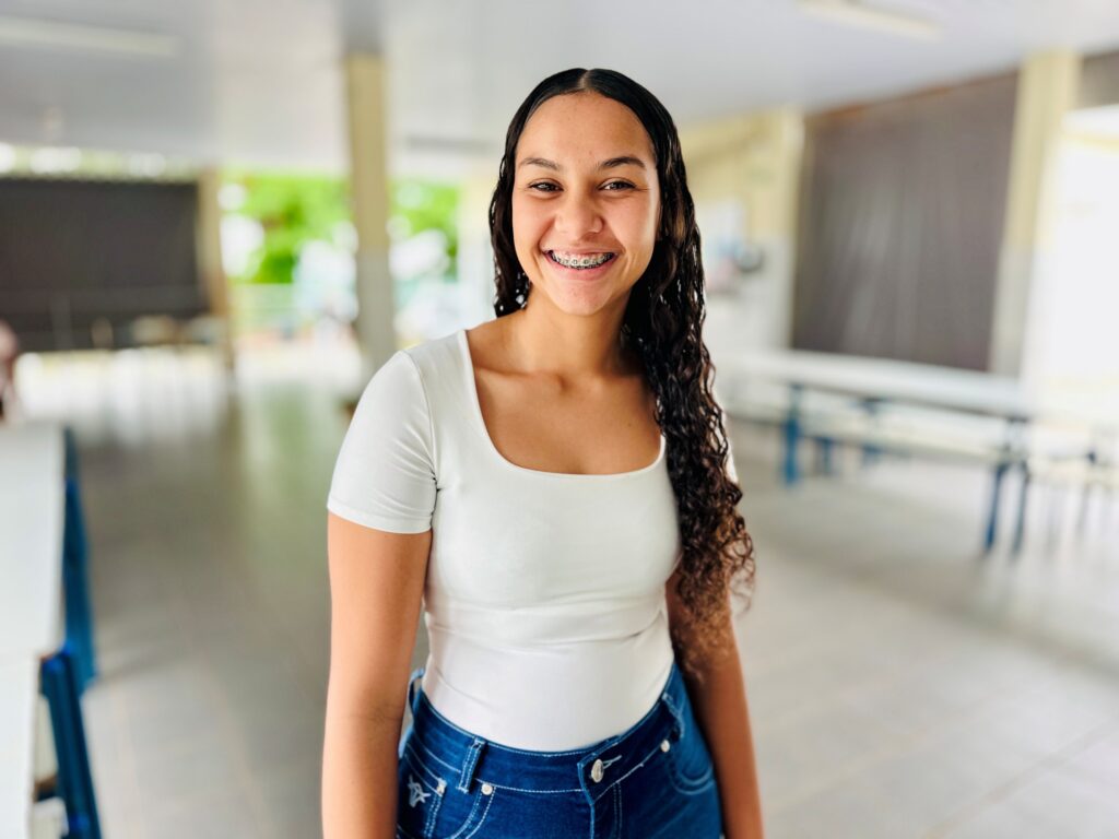 A imagem mostra uma jovem sorridente, com cabelos longos, escuros e cacheados. Ela está posicionada no centro da foto, que é um plano médio, e usa um sorriso que revela aparelho nos dentes. Sua pele é morena e seus olhos são escuros. Ela veste uma camiseta branca simples de decote redondo e uma calça jeans azul escura. O ambiente ao fundo parece ser um corredor ou área de convivência com pilares claros, piso de azulejos e algumas mesas e cadeiras ao fundo, tudo desfocado para destacar a pessoa na imagem. A iluminação é natural e clara, transmitindo uma sensação de alegria e leveza.