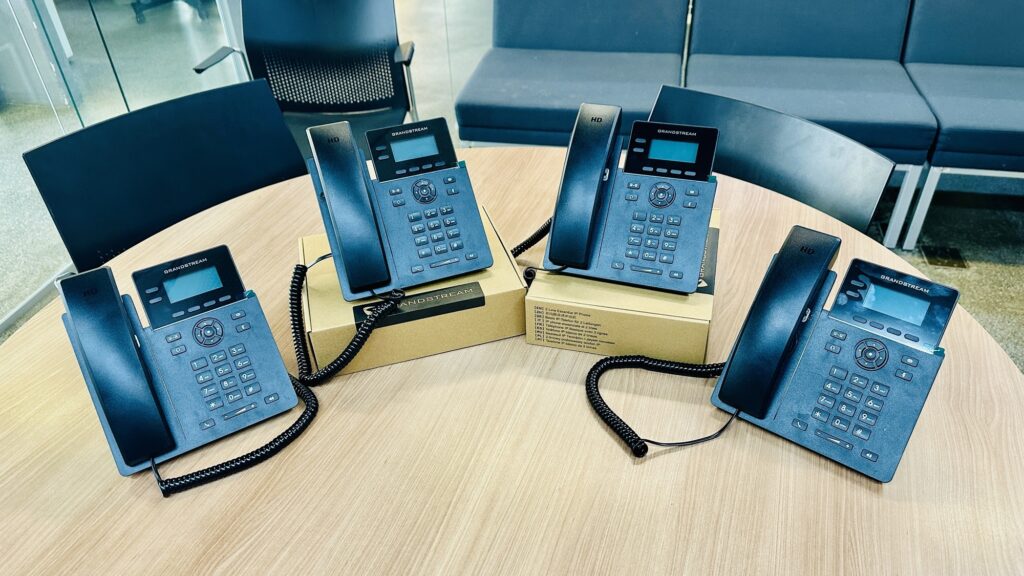 A imagem mostra uma mesa redonda de madeira clara com quatro telefones IP da marca Grandstream dispostos sobre ela. Dois dos telefones estão apoiados diretamente sobre a mesa, enquanto os outros dois estão em cima de caixas de papelão, provavelmente as embalagens originais dos aparelhos. Cada telefone é preto, com um pequeno visor retangular, teclas numéricas e botões de função, além do fone conectado por um cabo espiralado. O ambiente ao redor parece ser um escritório moderno, com cadeiras pretas e poltronas estofadas em azul ao fundo. A iluminação é clara e uniforme, destacando os detalhes dos telefones e da mesa. A imagem transmite uma sensação de organização e tecnologia, possivelmente relacionada à instalação ou apresentação de equipamentos de comunicação.