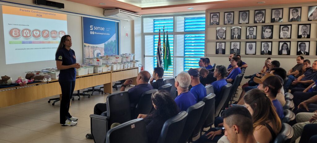 A imagem mostra uma sala de aula ou auditório onde está ocorrendo uma apresentação ou treinamento sobre Parada Cardiorrespiratória (PCR) e Suporte Básico de Vida (SBV). Na frente, uma mulher está de pé, provavelmente a instrutora, vestindo uma camisa polo azul escura com um logotipo e calça escura. Ao fundo, um projetor exibe um slide com o título "Parada Cardiorrespiratória PCR" e um gráfico intitulado "Figura 3. Cadeia de sobrevivência na PCR", além de informações sobre a "Atualização AHA 2025 Cadeia de Sobrevivência Unificada". Na mesa em frente à tela, há manequins para treinamento de RCP, tanto adulto quanto infantil, além de caixas que provavelmente contêm materiais para o curso. À direita, há um banner com o logotipo "Ssemae" e a frase "Um Novo Tempo", além de redes sociais (@semaepiracicaba). Os participantes estão sentados em cadeiras, assistindo atentamente à apresentação. A sala tem janelas grandes com persianas azuis e na parede há vários quadros com retratos de pessoas, possivelmente ex-diretores ou figuras importantes da instituição. Também é possível ver algumas bandeiras, incluindo a do Brasil. A atmosfera é de aprendizado e capacitação na área de emergência médica.
