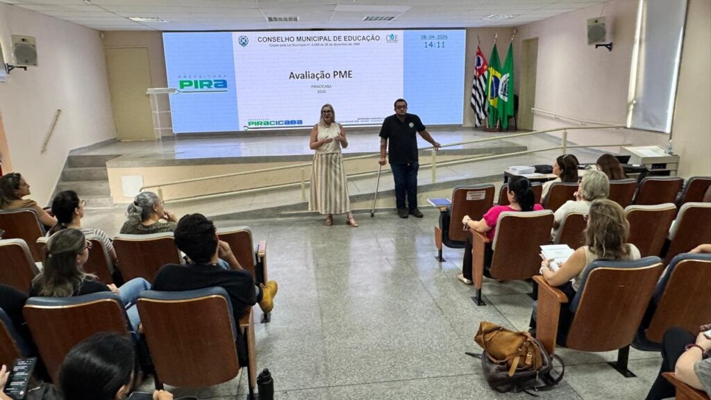 A imagem mostra uma reunião ou apresentação oficial acontecendo em um auditório. No centro, duas pessoas estão em pé diante de uma grande tela digital que exibe informações relacionadas ao "Conselho Municipal de Educação" de Piracicaba, com a data 08-04-2026 e a hora 14:11. A mulher, vestindo uma blusa clara e saia listrada, fala para a plateia, enquanto o homem ao lado usa uma muleta. Ao lado da tela, há três bandeiras, incluindo a do Brasil e a do estado de São Paulo. Na plateia, várias pessoas estão sentadas em cadeiras com encosto de madeira e estofado azul ou marrom, assistindo à apresentação. O ambiente é claro, com paredes claras, piso de azulejo e iluminação fluorescente no teto. Há também uma rampa com corrimão que leva ao palco onde os apresentadores estão. A atmosfera é formal e profissional, típica de uma reunião governamental ou educacional.