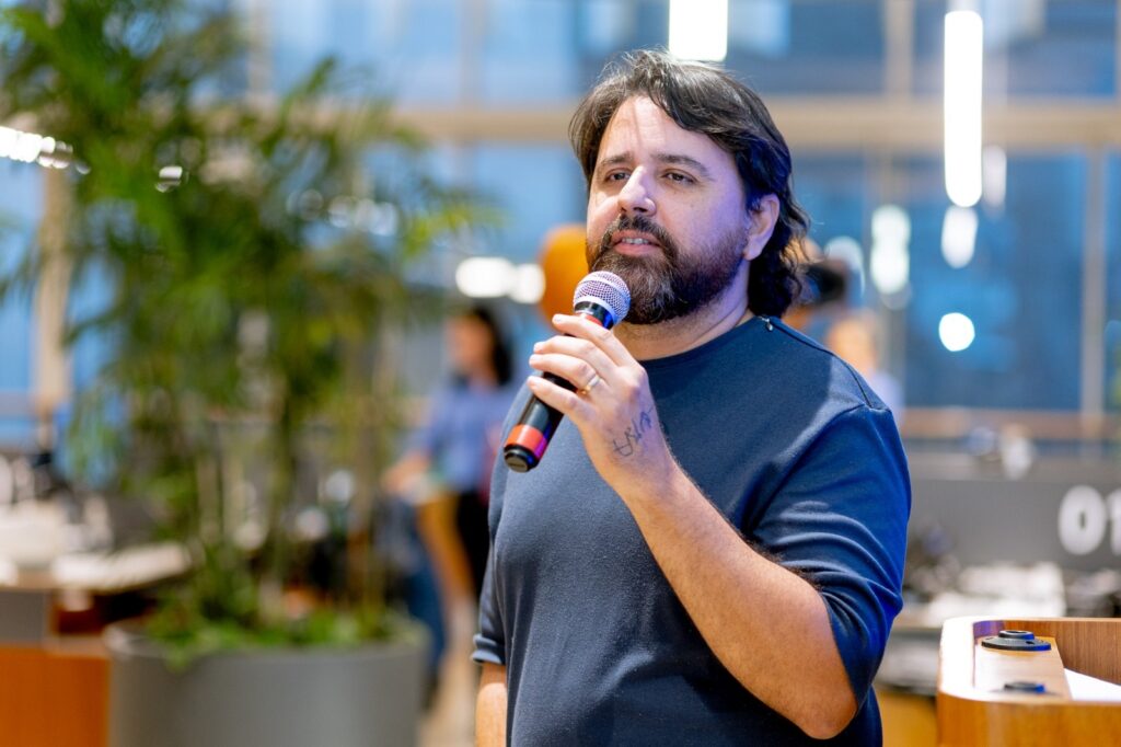 A imagem mostra um homem de meia-idade, com cabelos escuros e ondulados e barba cheia, falando ao microfone. Ele está vestido com uma camiseta azul-marinho e segura um microfone preto com a mão direita, onde se vê uma pequena tatuagem e uma aliança dourada no dedo anelar. O ambiente parece ser um espaço interno moderno, bem iluminado, com plantas grandes ao fundo e uma pessoa desfocada ao longe. A foto está focada no homem, enquanto o fundo está desfocado, destacando-o como o sujeito principal da imagem. À direita, há uma estrutura de madeira e o número "01" aparece parcialmente no fundo.