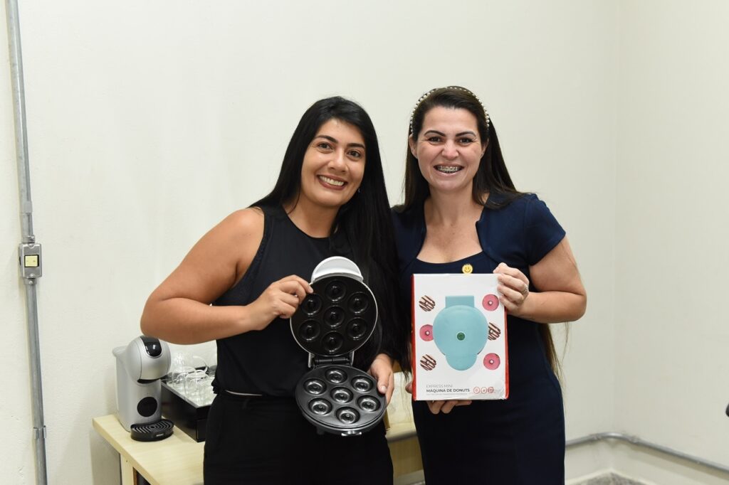 A imagem mostra duas mulheres sorridentes posando juntas em um ambiente interno, provavelmente um escritório ou sala de descanso. Elas estão atrás de uma mesa clara, sobre a qual está apoiada uma mini máquina de donuts preta aberta, exibindo sete cavidades para fazer donuts. Uma das mulheres segura a caixa do produto, que é branca com uma imagem da mini máquina de donuts em tom azul. Ao lado da máquina, há uma cafeteira prateada da marca Nescafé Dolce Gusto e algumas xícaras de vidro sobre uma bandeja. Ambas as mulheres têm cabelos longos e escuros; uma veste uma blusa preta sem mangas e a outra um vestido azul com uma tiara de pérolas. O ambiente tem paredes claras e iluminação uniforme, transmitindo uma atmosfera agradável e descontraída. A cena sugere um momento de celebração, presente ou promoção do produto “Express Mini Máquina de Donuts”.