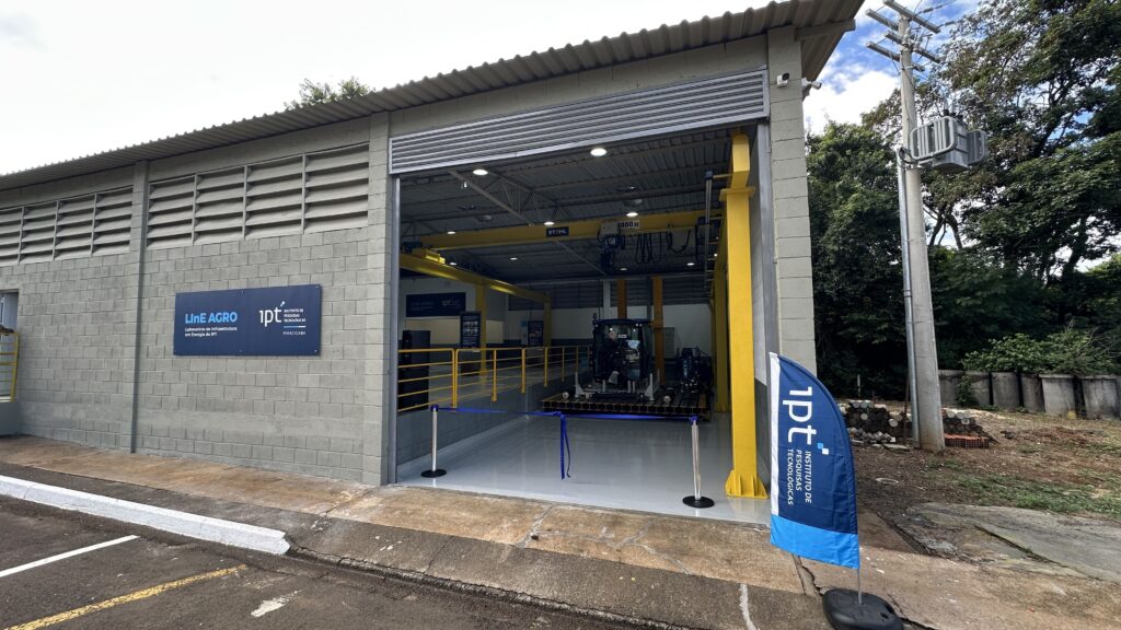 A imagem mostra a fachada e o interior de um laboratório industrial chamado "LinE AGRO", que pertence ao Instituto de Pesquisas Tecnológicas (IPT) em Piracicaba. O prédio é construído com blocos de concreto cinza e possui um telhado metálico ondulado. Na parte interna, há uma estrutura metálica amarela do tipo ponte rolante com a marca "STAHL" e capacidade de 3000 kg, indicando que é usada para movimentar equipamentos pesados. Dentro do laboratório, há uma máquina agrícola azul posicionada sobre uma plataforma. Na entrada, uma faixa azul delimita o espaço, sugerindo que o local está organizado para uma apresentação ou demonstração. Ao lado do prédio, há um poste com transformador, e ao fundo, árvores verdes indicam um ambiente externo bem cuidado. Na parede do laboratório, um grande letreiro azul exibe o nome "LinE AGRO Laboratório de Infraestrutura em Energia do IPT" e o logotipo do Instituto de Pesquisas Tecnológicas, reforçando o caráter tecnológico e de pesquisa do local. A imagem transmite a ideia de um espaço moderno dedicado ao desenvolvimento e estudo de tecnologias para o setor agroindustrial, com foco em infraestrutura energética.