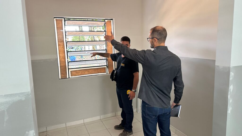 A imagem mostra dois homens dentro de uma sala vazia e simples, com paredes pintadas na metade inferior de cinza e na metade superior de branco, e piso de azulejos claros. Ambos estão de pé próximos a uma janela tipo veneziana, feita de várias lâminas horizontais de vidro. O homem mais próximo da câmera está de costas, veste uma camiseta cinza, calça jeans azul e usa óculos. Ele segura um caderno ou bloco de notas na mão esquerda e aponta com o braço direito em direção à janela. O outro homem, que está um pouco mais distante e de frente para o primeiro, usa uma camiseta preta com a inscrição "WSG" na frente, calça escura e uma bolsa de ombro preta. Ele também está gesticulando para a janela. A iluminação é clara e uniforme, provavelmente natural ou proveniente de luz artificial acima. A cena parece ser uma inspeção ou avaliação da janela ou do ambiente, sugerindo um contexto profissional, como construção, reforma ou vistoria imobiliária.