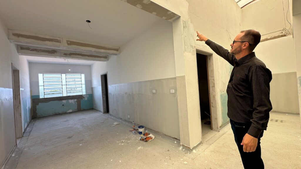 A imagem mostra um homem dentro de um ambiente que parece estar em reforma ou ainda não finalizado. Ele está em pé, virado para a esquerda, e aponta para cima com a mão direita, possivelmente indicando algo no teto. O homem usa óculos, tem cabelo curto e barba, e está vestido com uma camisa escura de manga longa com um padrão, além de calças escuras. O ambiente ao redor é simples e parece estar inacabado: o chão é de concreto e está coberto por poeira e resíduos. As paredes são claras, com uma faixa horizontal em tom cinza ou com reboco aparente, e algumas partes mostram sinais de desgaste ou acabamento incompleto. No teto, há tubulações expostas, reforçando a sensação de que o local está em processo de reforma. À esquerda, há uma janela com persianas horizontais, que deixam entrar a luz natural. No canto direito, próximo à parede, há alguns objetos no chão, incluindo um balde de tinta, ferramentas e uma garrafa plástica. A iluminação geral é natural, destacando o aspecto bruto do espaço. A cena transmite a ideia de inspeção ou avaliação do progresso da obra, com o homem apontando para algum detalhe no teto ou na estrutura.