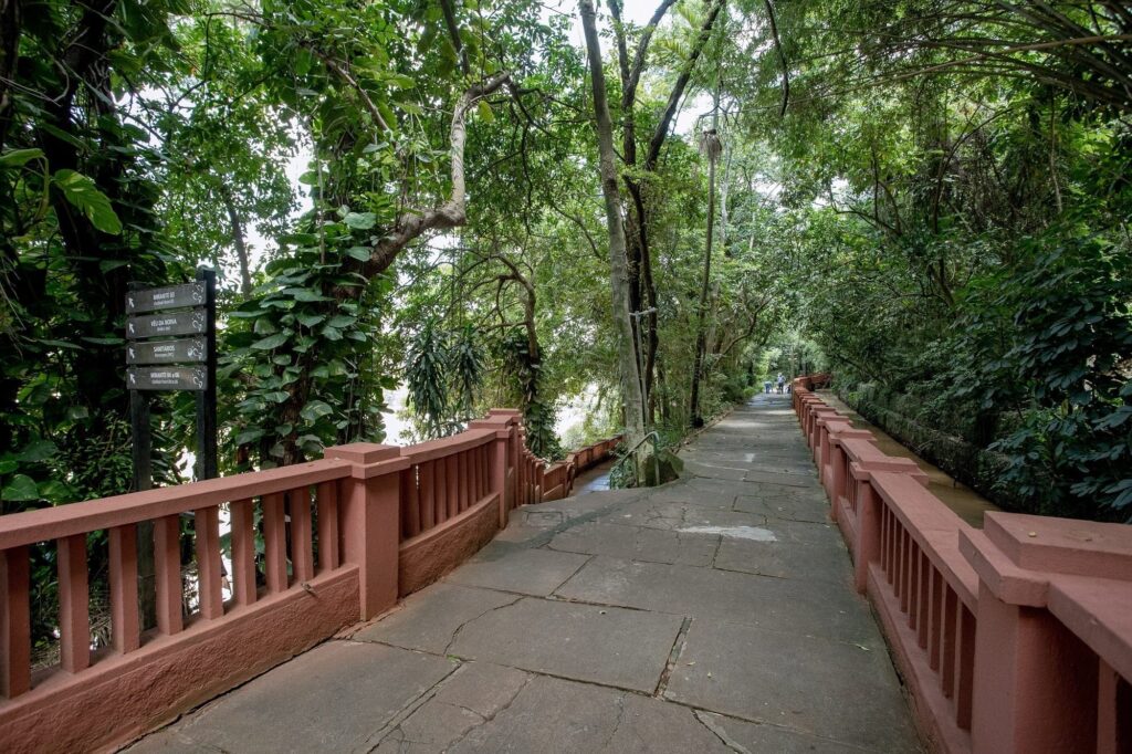 A imagem mostra um caminho pavimentado que serpenteia por uma floresta densa e verdejante. O caminho é feito de grandes pedras irregulares, algumas com musgo, indicando que é antigo e bastante usado. Ao longo do caminho, há um corrimão baixo de cor avermelhada que acompanha a curva do percurso e também desce por uma escadaria. À esquerda do caminho, há uma placa de sinalização com várias indicações em português, como “MIRANTE 03” (ponto de observação 03), “VÉU DA NOIVA”, “SANITÁRIOS” (banheiros) e “MIRANTE 04 a 06”. A placa está parcialmente coberta por trepadeiras, assim como as árvores ao redor, que são altas e com troncos grossos, formando uma copa densa que deixa passar luz em pontos, criando um efeito de luz e sombra no chão. O ambiente é natural e parece ser um parque ou jardim botânico, com muita vegetação e uma atmosfera tranquila. No fundo da imagem, é possível ver algumas pessoas caminhando, o que dá uma ideia da escala do local. A foto foi tirada de um ponto ligeiramente elevado, olhando para baixo, acompanhando a curva do caminho que desaparece na vegetação. A predominância de verdes e tons terrosos reforça o clima natural e acolhedor do lugar.