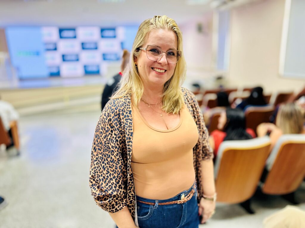A imagem mostra uma mulher loira sorridente em um ambiente que parece ser um auditório ou sala de aula. Ela está posicionada no centro-direito da foto, em foco nítido, enquanto o fundo está desfocado, mostrando fileiras de cadeiras de madeira e algumas pessoas sentadas de costas. A mulher usa óculos de armação transparente, uma blusa de tom bege claro com decote em "V", um kimono ou casaco com estampa de oncinha e jeans azul com um cinto fino marrom. Ela também usa um colar delicado e pequenos brincos. A iluminação é clara e uniforme, típica de ambientes internos profissionais ou educacionais. Ao fundo, à esquerda, há uma tela grande com um padrão quadriculado azul e branco, possivelmente um slide de apresentação ou logotipo. A composição da foto destaca a mulher com um sorriso acolhedor, transmitindo uma atmosfera amigável e descontraída.