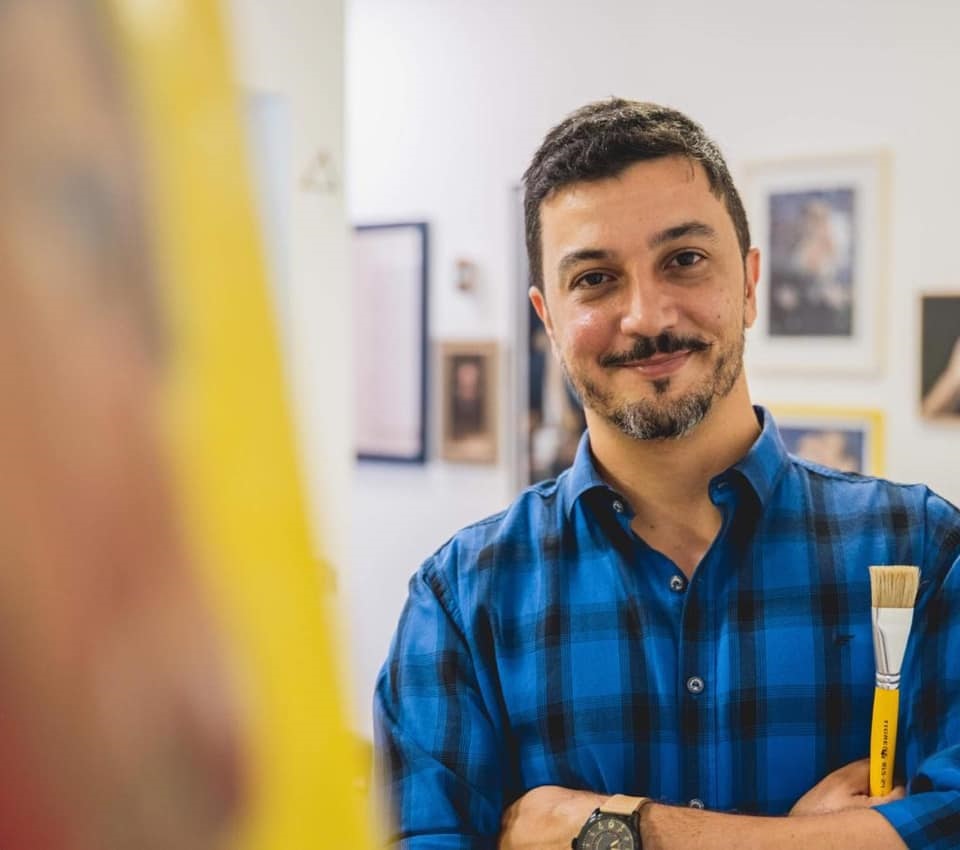 A imagem mostra um homem, provavelmente um artista, posando em um ambiente que parece ser um estúdio de arte ou uma galeria. Ele tem barba e bigode, usa uma camisa xadrez azul e preta, e está com os braços cruzados. Na mão direita, segura um pincel com cabo amarelo. O fundo é composto por várias molduras com quadros ou obras de arte penduradas na parede. A iluminação é suave, destacando o rosto do homem, que olha diretamente para a câmera com um leve sorriso. Em primeiro plano, do lado esquerdo, há um objeto amarelo desfocado, possivelmente parte de um cavalete ou tela. A composição sugere um momento de pausa e confiança do artista em seu espaço criativo.