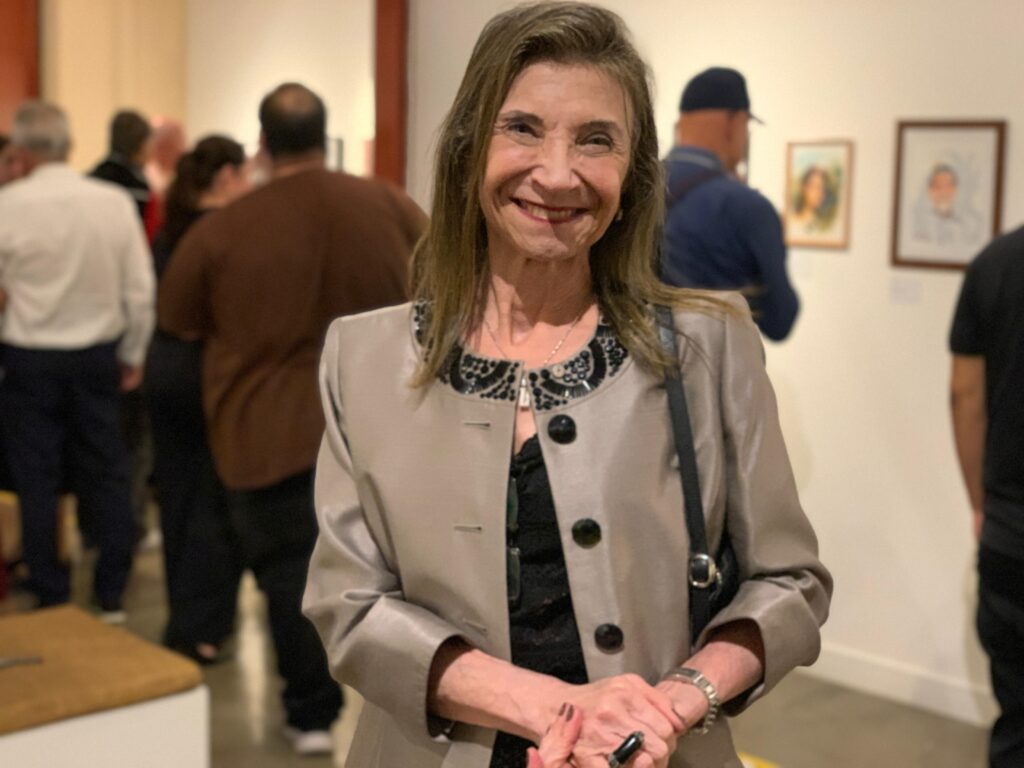 A imagem mostra uma mulher de meia-idade sorrindo, posando em um ambiente que parece ser uma galeria de arte ou museu. Ela está em foco nítido, enquanto o fundo está desfocado, com outras pessoas e quadros nas paredes, indicando que pode ser uma exposição pública. A mulher veste uma jaqueta clara, possivelmente de cetim, sobre uma blusa preta com um detalhe decorativo no decote. Ela usa um colar prateado com um pingente, um relógio no pulso esquerdo e um anel grande no dedo indicador direito. Suas mãos estão entrelaçadas à frente do corpo. A iluminação é suave e artificial, destacando seu rosto e parte superior do corpo. O contraste entre a jaqueta clara e a blusa escura chama atenção para a figura da mulher, enquanto o fundo desfocado sugere um ambiente cultural e social.