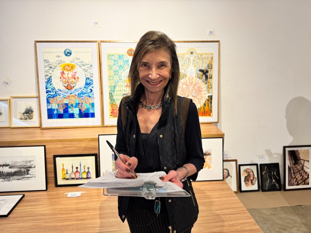 A imagem mostra uma mulher em uma galeria de arte, posicionada no centro da cena, olhando diretamente para a câmera com um sorriso amigável. Ela veste um colete preto acolchoado sobre um vestido preto e usa um colar chamativo com pedras quadradas transparentes. Em suas mãos, segura uma prancheta com papéis e uma caneta, sugerindo que está anotando ou revisando algo. Ao fundo, há várias obras de arte emolduradas expostas em prateleiras e penduradas na parede. As obras são variadas, incluindo peças coloridas e outras mais abstratas ou figurativas. Algumas etiquetas com números (como 13, 12, 36, 47) estão visíveis próximas às obras, provavelmente indicando números de catálogo ou preços. O ambiente é bem iluminado, com paredes brancas e prateleiras de madeira que criam um cenário limpo e minimalista, destacando as obras expostas. A perspectiva da foto é ao nível dos olhos, proporcionando uma visão clara e direta tanto da mulher quanto das artes ao seu redor. A cena sugere um evento de exposição ou venda de arte, com a mulher possivelmente atuando como artista, curadora ou responsável pela organização.