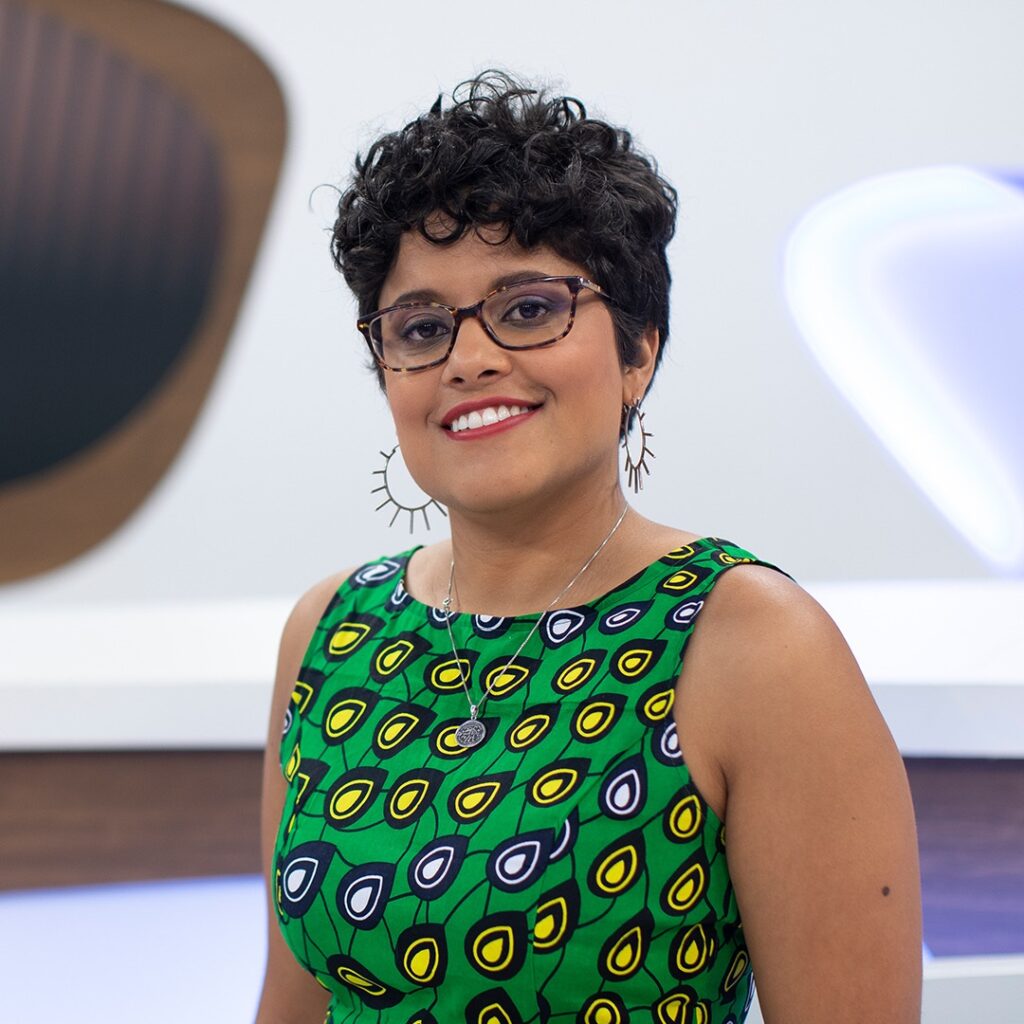 A imagem mostra uma mulher sorridente, de cabelo cacheado e escuro, usando óculos. Ela veste um vestido verde com estampa vibrante em tons de amarelo, branco e preto, com desenhos que lembram gotas. Ela também usa um colar prateado com um pingente redondo e brincos grandes em formato de sol. O fundo está desfocado, com uma iluminação que destaca seu rosto e roupa. A expressão dela é amigável e o ambiente parece ser um estúdio fotográfico ou de TV. A imagem transmite uma sensação de alegria e simpatia.