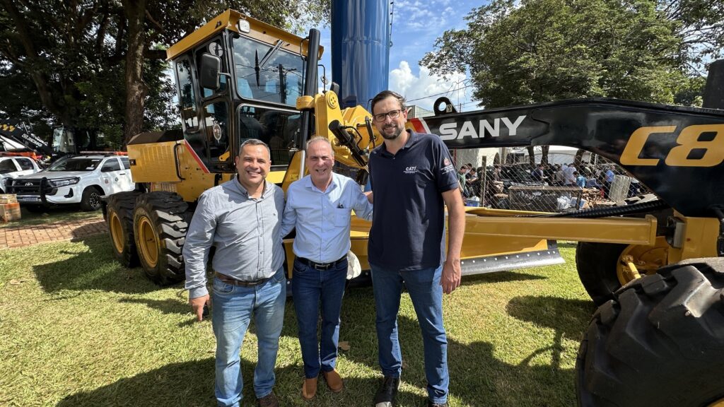 A imagem mostra três homens posando para a foto em frente a uma máquina pesada amarela, especificamente uma motoniveladora da marca SANY, modelo C8. Os três homens estão sorrindo e parecem estar em um ambiente externo, provavelmente em um evento ou apresentação da máquina. Dois dos homens vestem camisas sociais claras e jeans, enquanto o terceiro usa uma camisa polo azul escura com o logo da CATI (Coordenadoria de Assistência Técnica Integral) e do Governo do Estado de São Paulo, sugerindo uma ligação oficial ou institucional. Ao fundo, há outros veículos, incluindo um carro branco com placa visível, árvores e algumas pessoas atrás de uma cerca, o que indica que o local pode ser uma área aberta, como um pátio ou um campo de demonstração. A máquina, grande e robusta, domina a cena, destacando-se como o foco principal da imagem.