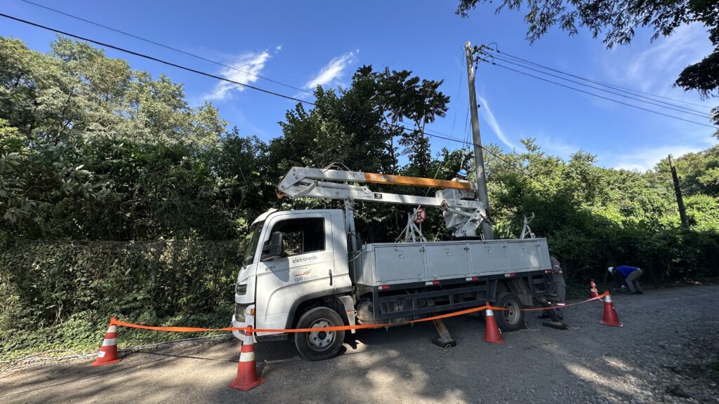 A imagem mostra uma cena ao ar livre em uma área rural ou semi-rural, com um caminhão utilitário branco da empresa "eletrorede" (parte da "cpfl paulista") estacionado em uma estrada de cascalho. O caminhão está equipado com um braço elevatório (guindaste) que está levantado para permitir que um trabalhador, usando capacete e uniforme azul, realize manutenção ou reparo em um poste de energia elétrica. O local ao redor do caminhão está isolado com cones de sinalização laranja e uma fita de segurança para evitar a passagem de pessoas. Ao fundo, há bastante vegetação verde e árvores, e o céu está azul com algumas nuvens brancas, indicando um dia claro. A cena transmite uma operação de manutenção de infraestrutura elétrica em andamento.