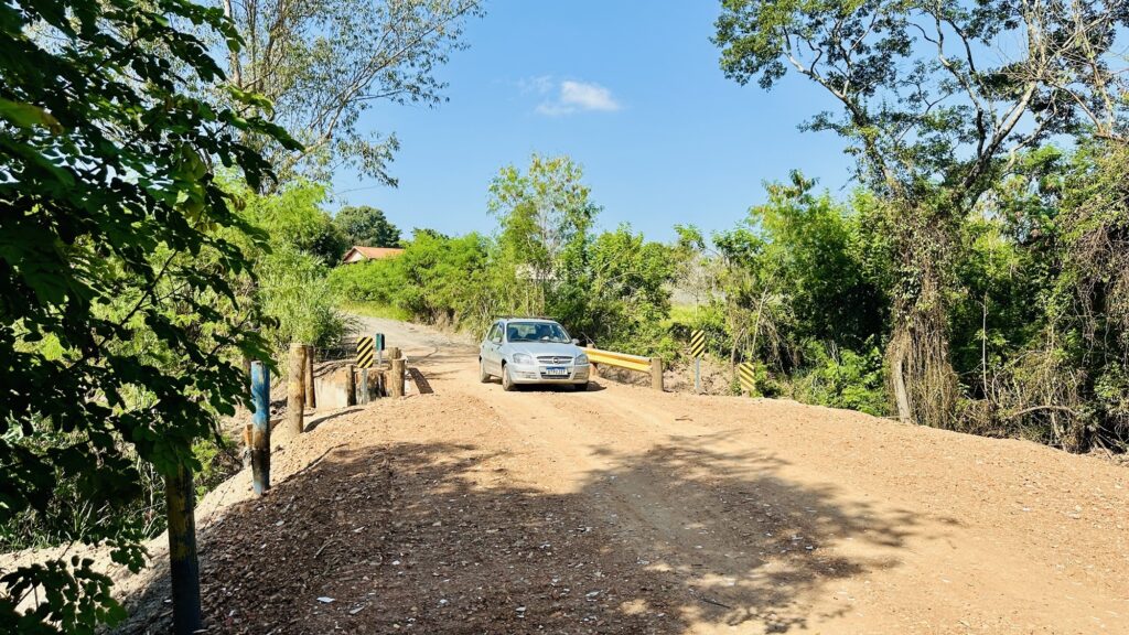 A imagem mostra um carro prata trafegando por uma estrada de terra que atravessa uma ponte. A estrada é de cascalho e terra, com uma superfície irregular. A ponte é feita de concreto, com postes de madeira e placas de sinalização amarelas e pretas em ambos os lados, indicando a presença da ponte. O carro está no centro da ponte, indo em direção ao observador. Ao redor da estrada, há uma vegetação verde e densa, com várias árvores. Ao fundo, é possível ver algumas casas com telhados vermelhos, parcialmente escondidas entre as árvores. O céu está claro, azul, com poucas nuvens brancas, e a iluminação sugere que é um dia ensolarado. A cena transmite uma sensação de área rural tranquila e natural.