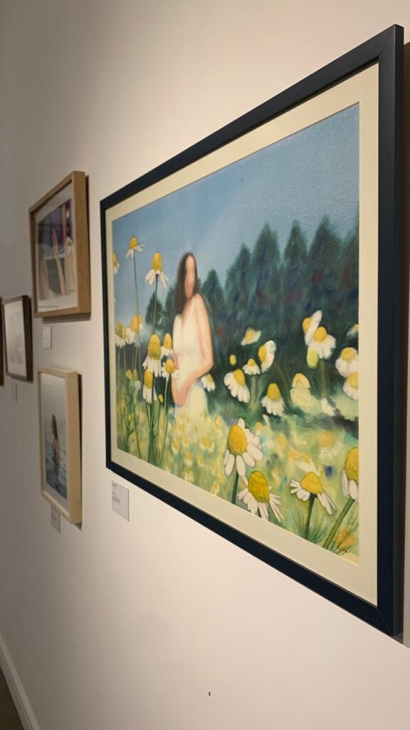A imagem mostra uma pintura emoldurada pendurada em uma parede branca, provavelmente em uma galeria de arte. A pintura retrata uma mulher em pé em um campo de margaridas. A mulher está com o rosto desfocado, vestindo um vestido claro, e segurando as mãos à frente do corpo. As margaridas são grandes e destacadas, com pétalas brancas e centros amarelos, espalhadas por um campo em tons de verde e amarelo. Ao fundo, há um grupo denso de árvores verdes escuras sob um céu azul claro. A pintura tem um estilo que mistura impressionismo com um toque onírico, devido ao rosto desfocado da mulher e à suavidade das cores e formas. A obra está emoldurada com uma moldura escura e uma borda azul escura ao redor da pintura. Ao lado, à esquerda, há outras pinturas penduradas na parede, em molduras diferentes, mostrando cenas variadas, mas estão menos em destaque. A iluminação é uniforme, típica de galerias, destacando as obras na parede. A foto foi tirada de um ângulo ligeiramente baixo, olhando para cima.