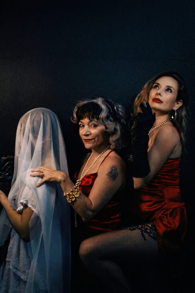 A imagem mostra um retrato dramático e artístico de três mulheres contra um fundo escuro e texturizado. As três estão vestidas com vestidos vermelhos de cetim, criando um forte contraste com o fundo preto. A mulher do centro tem cabelos grisalhos e usa um colar de pérolas, pulseiras douradas e um vestido vermelho, com um sorriso confiante e olhar direto para a câmera. À esquerda, uma mulher está parcialmente coberta por um véu branco translúcido, sugerindo um tema de mistério ou noiva, com apenas parte do rosto e braço visíveis. À direita, outra mulher loira usa luvas longas pretas e meia arrastão, olhando para cima com uma expressão pensativa ou dramática. A iluminação é forte e direcionada, destacando os rostos e as texturas dos vestidos e acessórios, enquanto o fundo escuro cria um clima intenso e teatral. A composição transmite uma atmosfera de mistério, elegância e um toque de teatralidade.