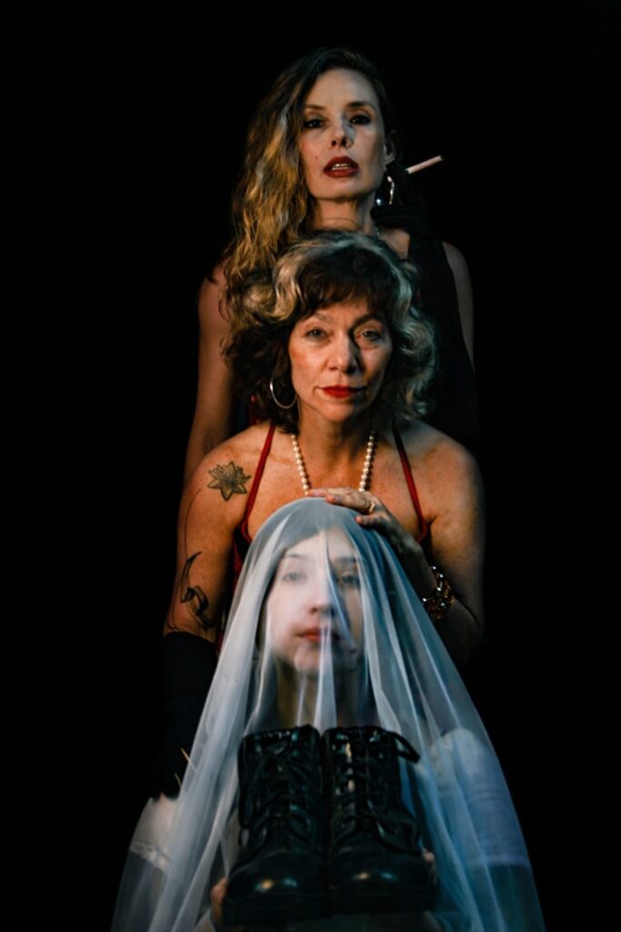 A imagem mostra um retrato em plano médio de três mulheres dispostas em camadas verticais contra um fundo preto. Na frente, uma mulher está parcialmente coberta por um véu branco translúcido, com seu rosto suavemente iluminado e olhando diretamente para a câmera. Ela segura um par de botas pretas de combate à sua frente. Atrás dela, uma segunda mulher, com cabelo cacheado e mechas claras, usa um colar de pérolas e uma blusa vermelha; suas mãos repousam sobre o véu da mulher da frente e seu olhar é sério, direcionado para frente. A terceira mulher está em pé ao fundo, com cabelo longo e ondulado, segurando um cigarro aceso na boca e olhando fixamente para a câmera, exibindo uma expressão severa. A iluminação dramática destaca os rostos e cria forte contraste com o fundo escuro, conferindo à cena um clima intenso e carregado de simbolismo, reforçado pela presença do véu e das botas de combate.