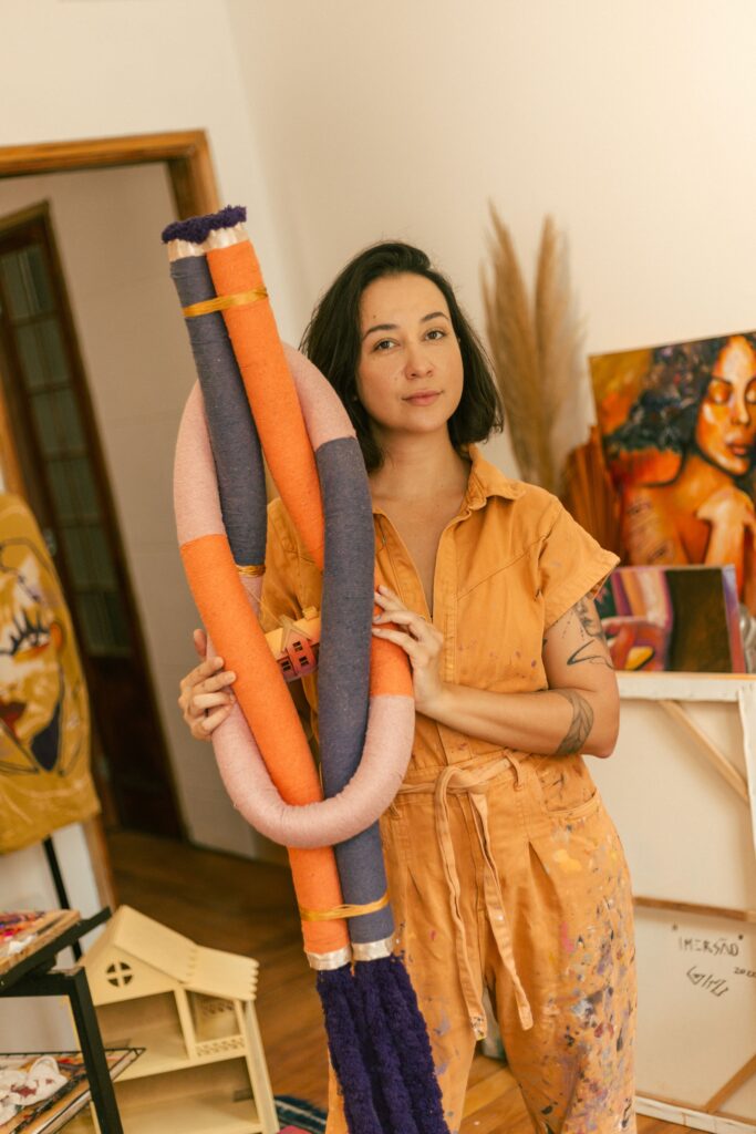 A imagem mostra uma mulher em um estúdio de arte, segurando uma escultura grande e colorida que tem formato cilíndrico e é composta por tubos entrelaçados em diferentes cores e texturas, como laranja, rosa, azul escuro e roxo, amarrados com fitas douradas. A mulher, que parece ser a artista, está vestindo um macacão laranja com manchas de tinta, sugerindo que está no meio do processo criativo. Ela olha diretamente para a câmera com uma expressão neutra. Ao fundo, o ambiente é claramente um ateliê, com várias obras de arte visíveis, incluindo uma pintura de rosto em um cavalete e outra obra com um rosto estilizado em outra parte do estúdio. Também há uma pequena estante que se assemelha a uma casinha de bonecas. A iluminação é suave e natural, criando uma atmosfera acolhedora e artística. Na parte superior da imagem, há o texto "IMERSAO LIVIA 2022", possivelmente indicando o nome da exposição, do projeto ou da artista, e o ano. A composição da foto destaca tanto a artista quanto a sua obra, mostrando o ambiente de criação e dando um contexto ao trabalho apresentado.