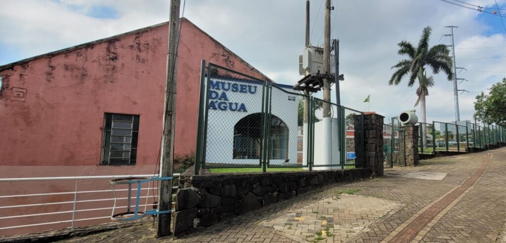 A imagem mostra a entrada do Museu da Água, localizada em uma área urbana com um céu parcialmente nublado ao fundo. À esquerda, há um prédio de cor rosa claro com uma janela pequena, cuja pintura está um pouco desgastada. Em frente ao museu, há uma cerca de metal verde que delimita a entrada. A parede do museu é branca, com o nome "MUSEU DA ÁGUA" escrito em letras grandes e azuis. Ao lado, há um poste com um transformador elétrico e fios que se estendem pela imagem. No canto direito, vê-se uma parede feita de pedras, um calçamento de pedras irregulares e uma palmeira alta. O ambiente parece tranquilo, sem pessoas visíveis, e a imagem é capturada a partir de um ângulo baixo, mostrando uma leve inclinação da rua. Ao fundo, também é possível notar a presença da bandeira do Brasil, sugerindo que o museu está localizado no país.