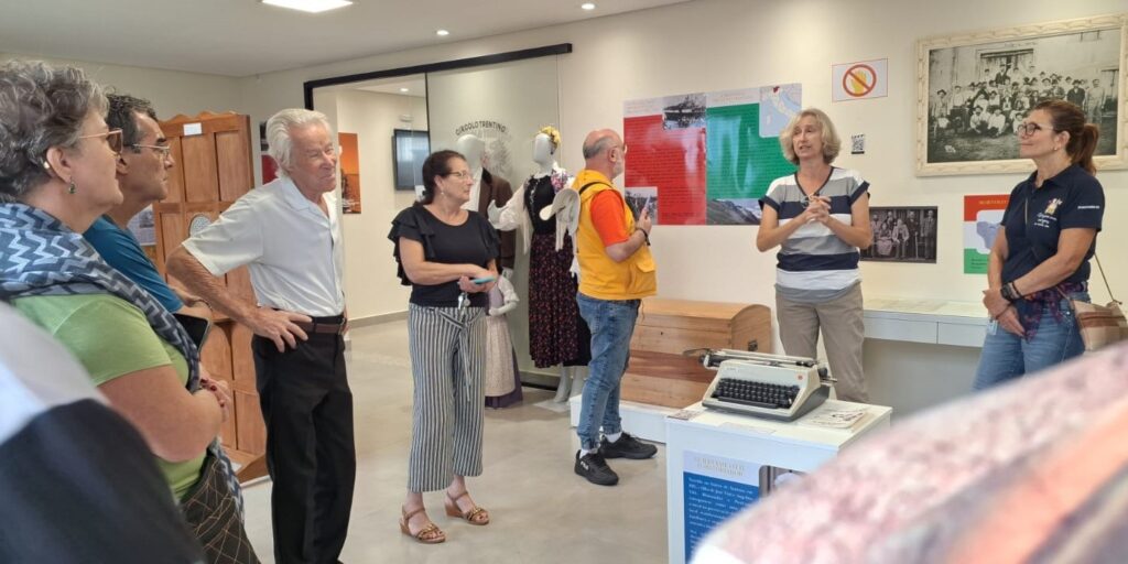 A imagem mostra um grupo de pessoas reunidas em um ambiente interno, que parece ser um centro cultural ou museu dedicado à história e à cultura italiana, possivelmente ligado à imigração italiana no Brasil, dado o nome "Circolo Trentino" visível na porta de vidro. Ao centro da cena, uma mulher está falando e gesticulando para o grupo, que a escuta atentamente. Ela está perto de um pedestal branco onde há uma máquina de escrever antiga e, ao lado, um baú de madeira. Ao fundo, há manequins vestidos com trajes folclóricos tradicionais italianos, além de painéis e fotografias históricas expostas nas paredes. O ambiente é bem iluminado, com luzes embutidas no teto, e as pessoas presentes são de várias idades, com destaque para um homem idoso vestindo uma camisa pólo branca e uma mulher com blusa preta e calça listrada. Na parede ao fundo, há um cartaz com as cores da bandeira italiana (verde, branco e vermelho) e textos informativos. No geral, a imagem transmite uma atmosfera de aprendizado e valorização da cultura e história italiana, com um grupo engajado em uma palestra ou visita guiada.